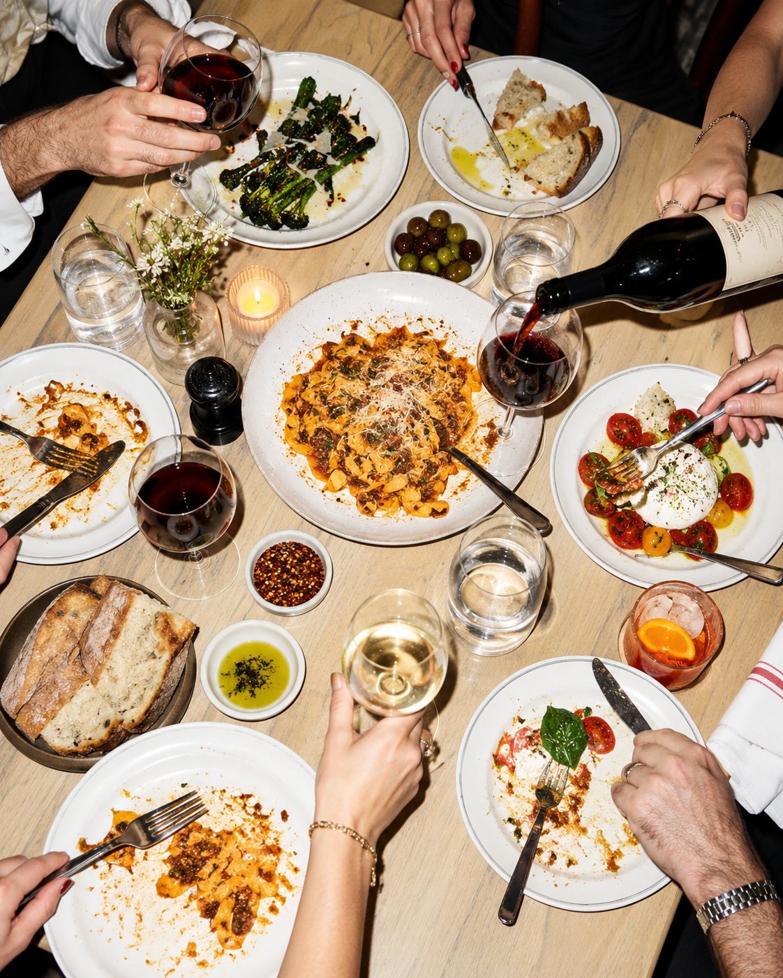 Seen from above, a shared dinner mid-meal: red wine being poured, pasta and burrata half eaten, bread torn, and hands reaching across the table